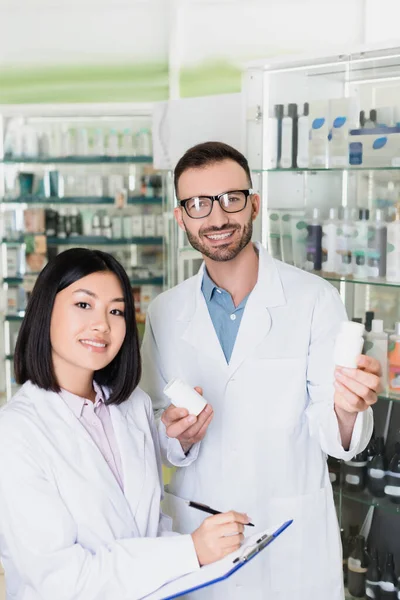 Lächelnder Apotheker im weißen Kittel mit Flaschen mit Medikamenten in der Nähe eines asiatischen Kollegen in der Apotheke — Stockfoto