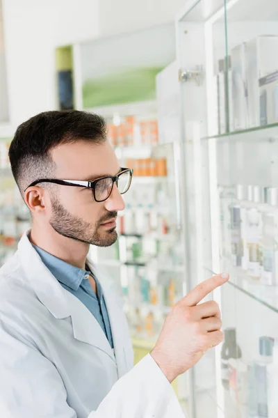 Farmacéutico en bata blanca y anteojos apuntando con el dedo en la farmacia - foto de stock