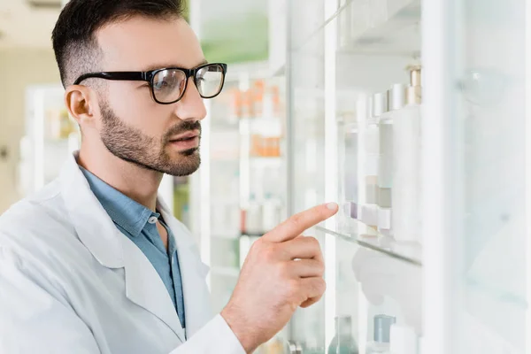 Farmacéutico barbudo con abrigo blanco y anteojos apuntando con el dedo en la farmacia - foto de stock