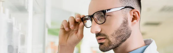 Bearded pharmacist adjusting eyeglasses in drugstore, banner — Stock Photo