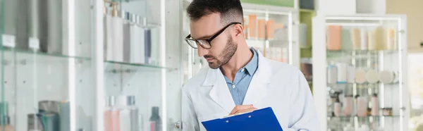 Farmacéutico barbudo con abrigo blanco y anteojos sujetando el portapapeles mientras revisa la medicación en la farmacia, pancarta - foto de stock