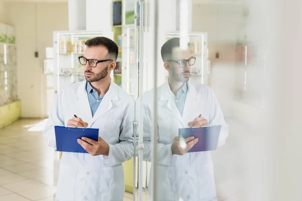 Bearded pharmacist in eyeglasses holding clipboard while checking medication in drugstore — Stock Photo