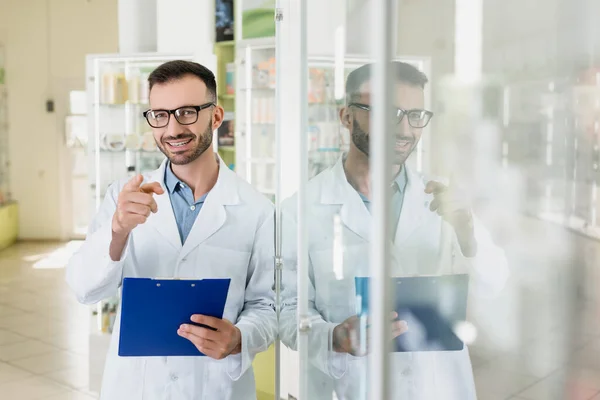 Pharmacien heureux dans les lunettes tenant presse-papiers tout en pointant du doigt dans la pharmacie — Photo de stock