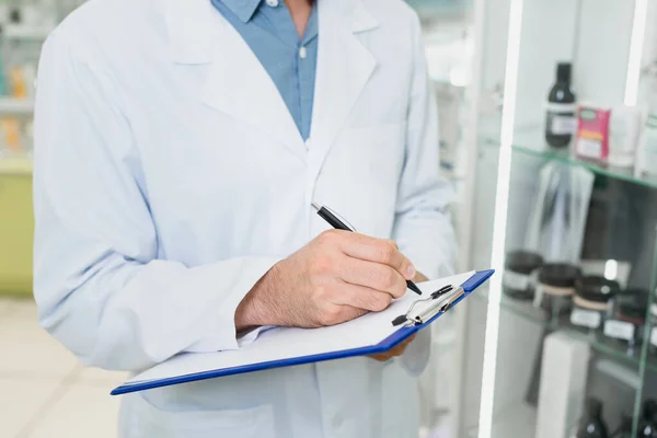 Partial view of pharmacist writing on clipboard in drugstore — Stock Photo