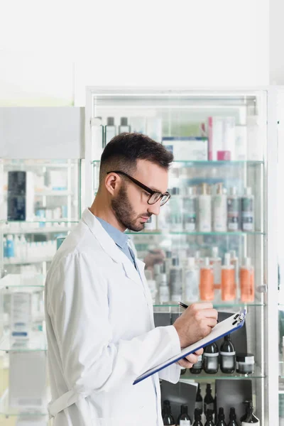 Farmacéutico barbudo en anteojos escribiendo en portapapeles en farmacia - foto de stock