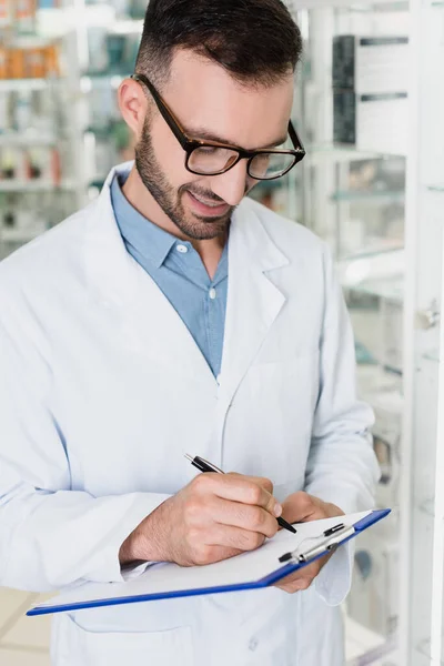 Farmacéutico alegre en las gafas que escriben en portapapeles en la farmacia - foto de stock