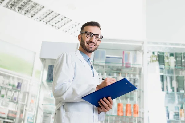 Cheerful pharmacist in eyeglasses and white coat writing on clipboard in drugstore — Stock Photo