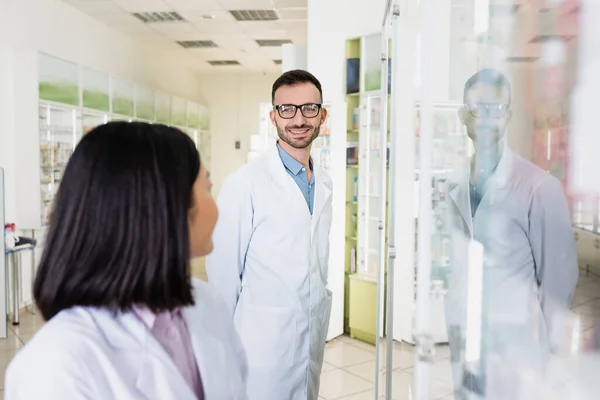 Cheerful pharmacist in eyeglasses and white coat looking at brunette asian colleague on blurred foreground — Stock Photo