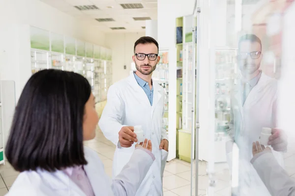 Bärtiger Apotheker mit Brille und weißem Mantel gibt Flasche mit Pillen an brünetten asiatischen Kollegen auf verschwommenem Vordergrund — Stockfoto