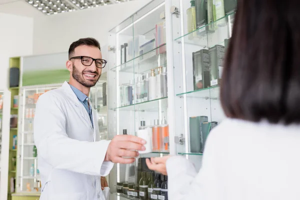 Heureux pharmacien dans des lunettes et manteau blanc donnant bouteille avec des pilules à collègue brune sur le premier plan flou — Photo de stock