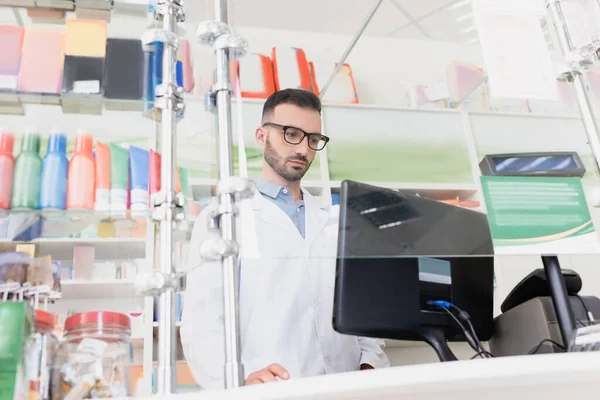 Pharmacien en manteau blanc et lunettes regardant moniteur d'ordinateur en pharmacie — Photo de stock