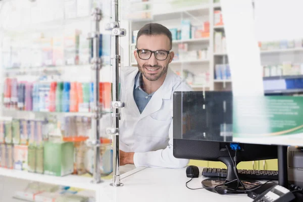 Farmacêutico feliz em casaco branco e óculos olhando para a câmera perto do monitor do computador na farmácia — Fotografia de Stock