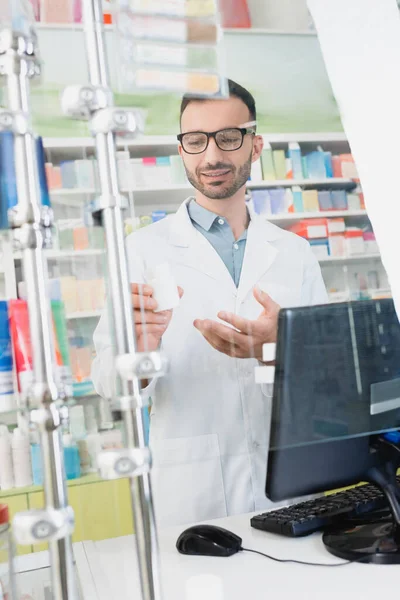 Happy pharmacist in white coat and eyeglasses pointing with hand at bottle near computer monitor in drugstore — Stock Photo