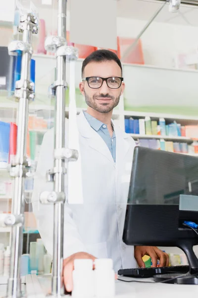 Smiling pharmacist in white coat and eyeglasses looking at camera near computer monitor in drugstore — Stock Photo