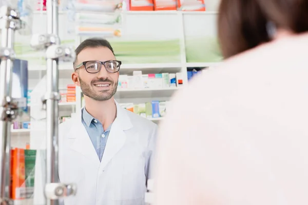 Smiling pharmacist in white coat and eyeglasses looking at customer on blurred foreground — Stock Photo