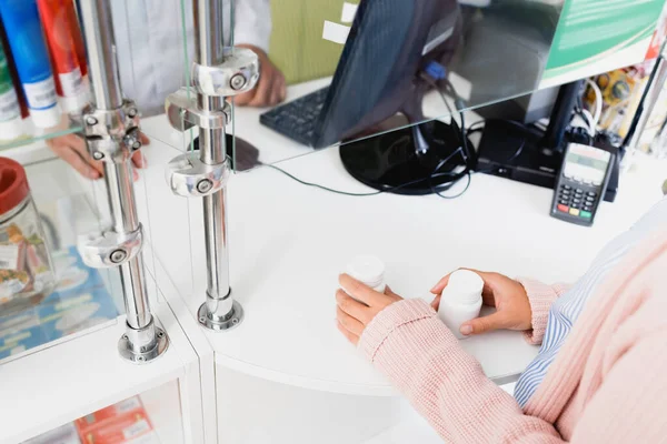 Vista recortada de los clientes sosteniendo botellas con pastillas cerca del vendedor en la farmacia - foto de stock