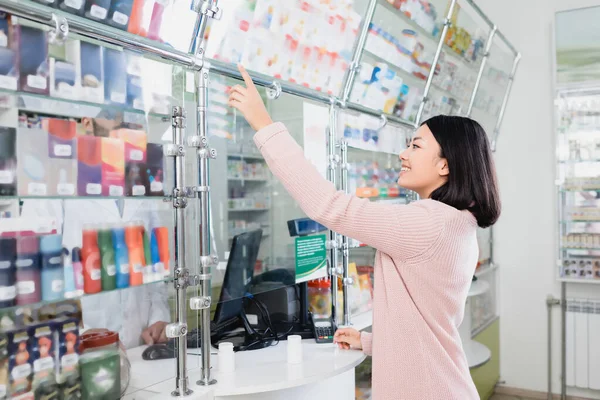 Feliz asiático cliente apuntando con el dedo en la medicación cerca vendedor en droguería - foto de stock