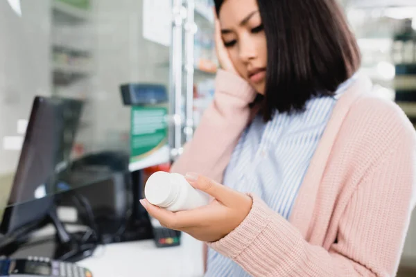 Asiatin hält Flasche mit Tabletten, während sie Kopfschmerzen in Drogerie hat — Stockfoto