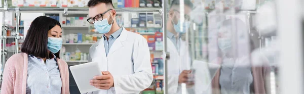 Pharmacien en lunettes et masque médical tenant comprimé numérique près de la femme asiatique en pharmacie, bannière — Photo de stock