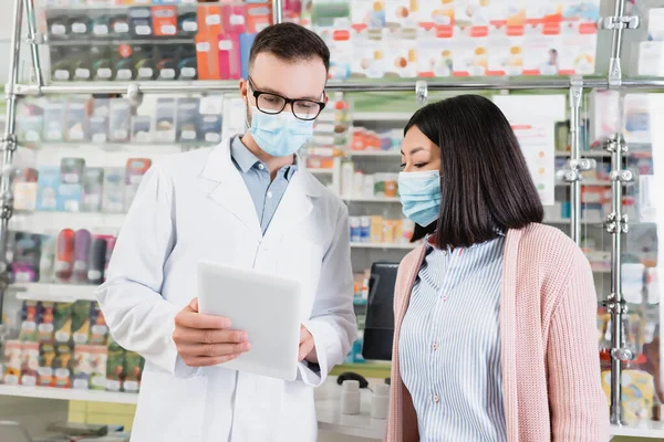 Pharmacien en lunettes et masque médical tenant comprimé numérique près de la femme asiatique en pharmacie — Photo de stock