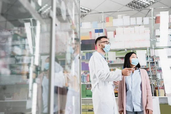 Apotheker in medizinischer Maske schaut mit asiatischer Kundin in Drogerie weg — Stockfoto