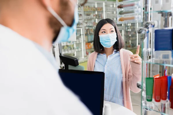 Asiático cliente no médico máscara apontando com dedo perto farmacêutico no embaçado primeiro plano — Fotografia de Stock