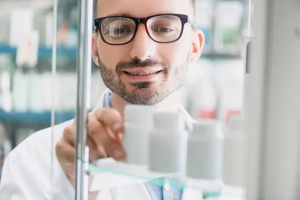 Pharmacien joyeux dans des lunettes de vue prenant bouteille avec des pilules de l'étagère en verre sur le premier plan flou — Photo de stock