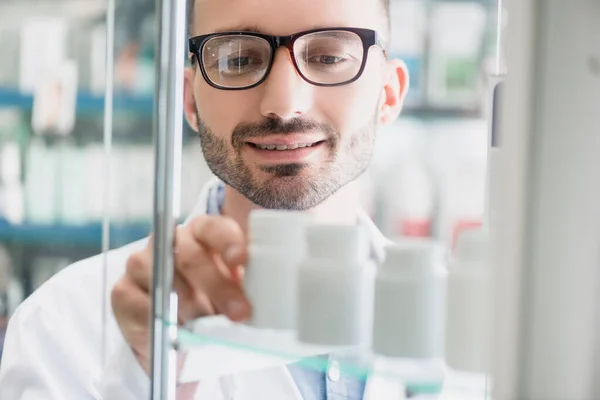 Gut gelaunter Apotheker in Brille nimmt Flasche mit Pillen aus Glasregal im verschwommenen Vordergrund — Stockfoto