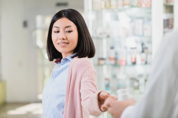 Alegre asiático mujer tomando botella con píldoras cerca de farmacéutico en borrosa primer plano - foto de stock