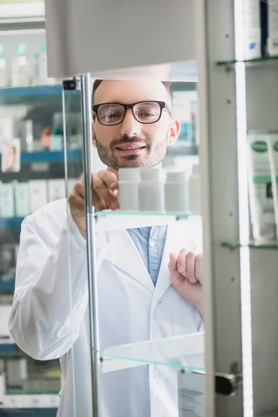 Farmacêutico feliz em óculos tomando garrafa com pílulas de prateleira de vidro em primeiro plano borrado — Fotografia de Stock