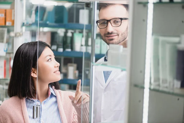 Zufriedene asiatische Kundin zeigt mit Finger auf bärtigen Apotheker in Drogerie — Stockfoto