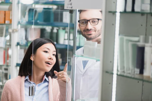 Curioso asiático cliente apuntando con dedo cerca barbudo farmacéutico en droguería - foto de stock