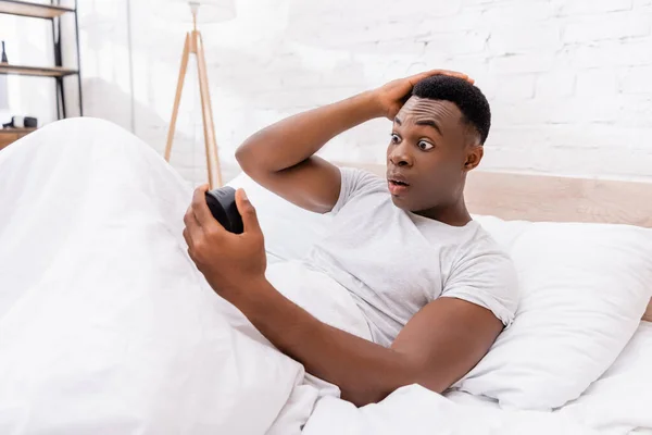 Aufgeregter afrikanisch-amerikanischer Mann schaut morgens auf die Uhr im Bett — Stockfoto