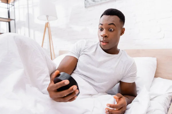 Homme afro-américain excité regardant l'horloge sur le premier plan flou sur le lit le matin — Photo de stock