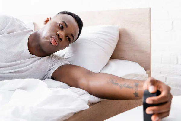 Hombre afroamericano haciendo pucheros labios mientras mira el reloj en primer plano borroso cerca de la cama - foto de stock