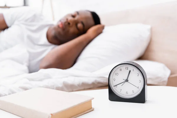Clock and book on bedside table near african american man sleeping on blurred background — Stock Photo