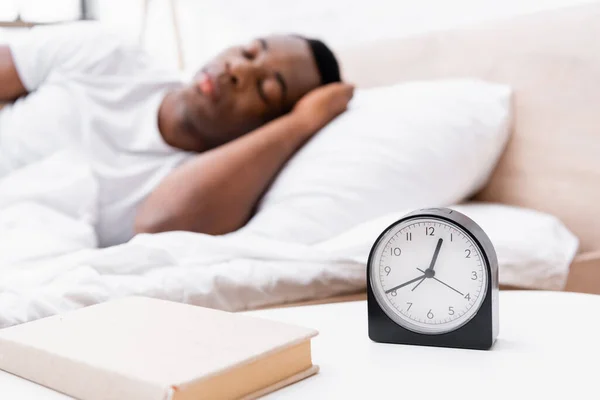 Orologio e libro sul comodino vicino all'uomo afroamericano che dorme su sfondo sfocato — Foto stock