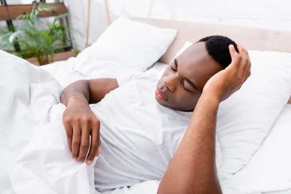 Hombre afroamericano tocando la cabeza mientras está acostado en la cama - foto de stock