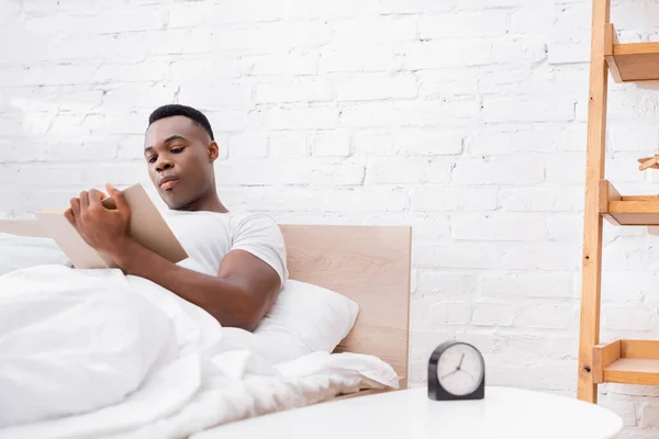 Afrikanischer Mann liest Buch nahe Uhr auf Nachttisch auf verschwommenem Vordergrund — Stockfoto