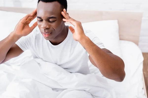Afrikanischer Mann mit geschlossenen Augen leidet unter Kopfschmerzen im Bett vor verschwommenem Hintergrund — Stockfoto