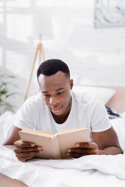 Africano americano homem lendo livro enquanto deitado na cama de manhã — Fotografia de Stock