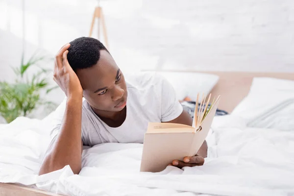 Hombre afroamericano enfocado leyendo libro en la cama en casa - foto de stock