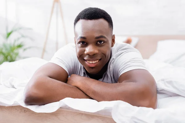 Homme afro-américain joyeux regardant la caméra tout en étant couché sur le lit à la maison — Photo de stock