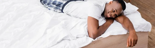 African american man in pajamas smiling while lying on bed, banner — Stock Photo