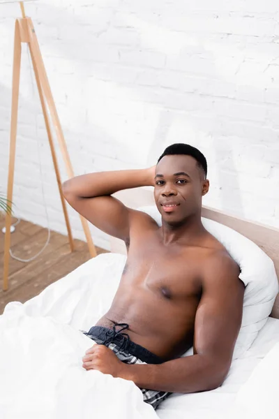 Musculoso afroamericano hombre sonriendo a la cámara en la cama en casa - foto de stock
