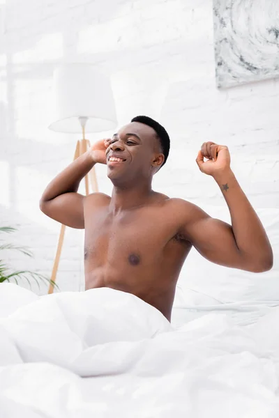 Shirtless afroamericano hombre sonriendo y estirándose en la cama - foto de stock