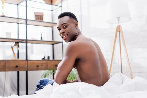 Shirtless african american man smiling at camera on white bedding at home — Stock Photo