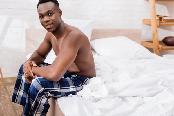 Sans chemise homme afro-américain assis sur le lit et souriant à la caméra — Photo de stock