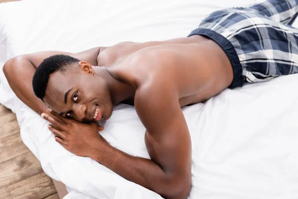 Hemdloser afrikanisch-amerikanischer Mann lächelt in die Kamera, während er auf dem Bett liegt — Stockfoto