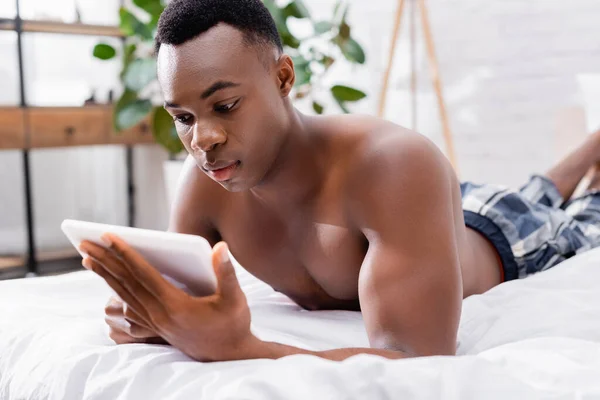 Африканский мужчина без рубашки использует цифровые планшеты на размытом переднем плане на кровати — стоковое фото